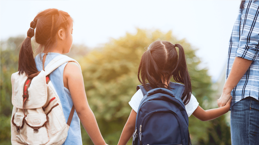Two girls with backpacks walk towards school with father